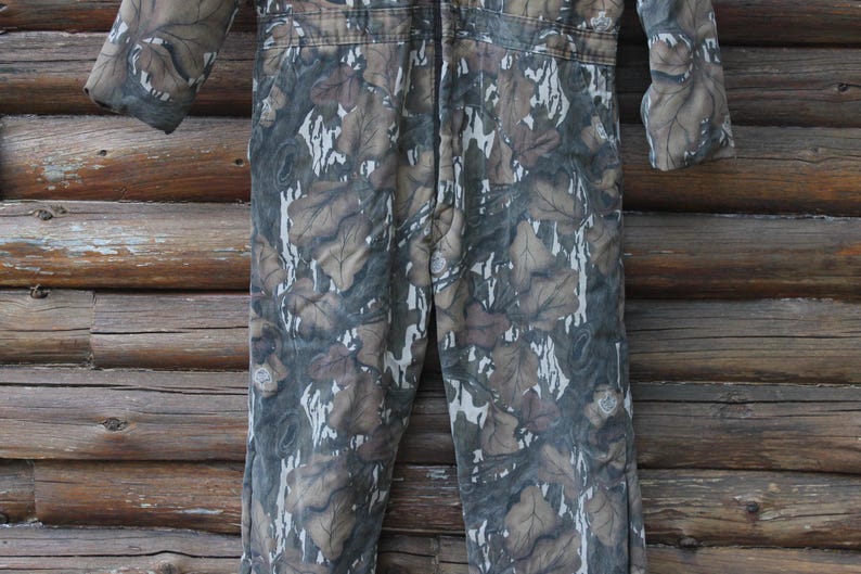 Puede incluir: Mono con estampado de camuflaje y cierre de cremallera. La prenda de una pieza presenta un dise&ntilde;o de hojas marrones, verdes y blancas. El mono de manga larga est&aacute; hecho de una tela duradera, adecuada para actividades al aire libre.