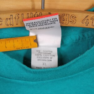 May include: Teal blue t-shirt with a white tag that reads "Made Exclusively for The Walt Disney Theme Parks and Resorts". The tag also has the text "Disney" on it. The t-shirt has a size tag that reads "XL" and "100% Cotton Exclusive of Decoration".