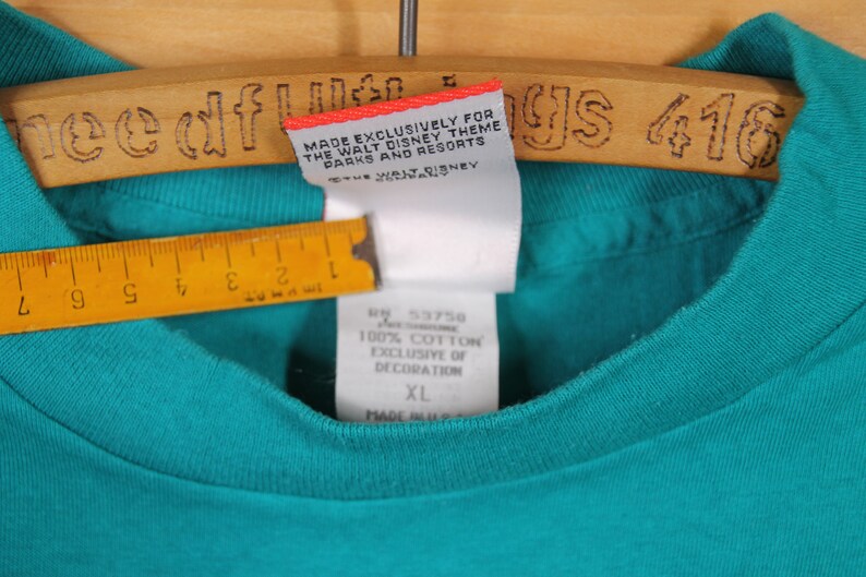 May include: Teal blue t-shirt with a white tag that reads "Made Exclusively for The Walt Disney Theme Parks and Resorts". The tag also has the text "Disney" on it. The t-shirt has a size tag that reads "XL" and "100% Cotton Exclusive of Decoration".