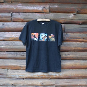 Op de afbeelding: Zwart t-shirt met drie kleurrijke vierkante afbeeldingen. De afbeeldingen lijken illustraties te zijn van figuren in actie. Het shirt hangt aan een houten hanger tegen een houten achtergrond.