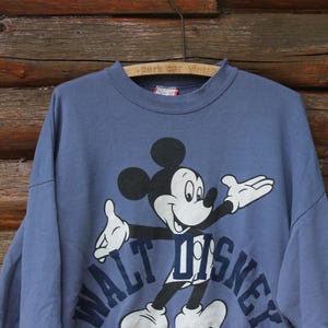 Puede incluir: Sudadera azul vintage con un gr&aacute;fico de Mickey Mouse. Las palabras "WALT DISNEY" est&aacute;n impresas en azul marino debajo del personaje. La sudadera est&aacute; colgada de una percha de madera.