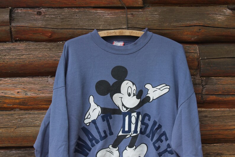 Puede incluir: Sudadera azul vintage con un gr&aacute;fico de Mickey Mouse. Las palabras "WALT DISNEY" est&aacute;n impresas en azul marino debajo del personaje. La sudadera est&aacute; colgada de una percha de madera.