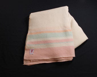 Op de afbeelding: Een gevouwen crèmekleurige deken met horizontale strepen in perzik, groen en roze. Een klein label is zichtbaar in de onderste hoek. De deken is gemaakt van een zacht materiaal en is netjes opgevouwen.