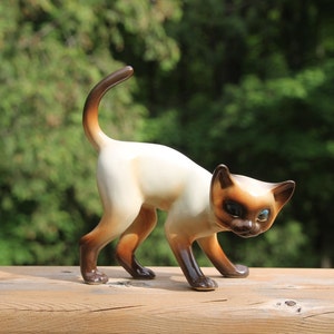 Op de afbeelding: Een keramiek beeldje van een Siamese kat met bruin en wit bont. De kat zit in een hurkende houding met zijn staart opgerold. De kat heeft blauwe ogen en een witte vlek op zijn gezicht.