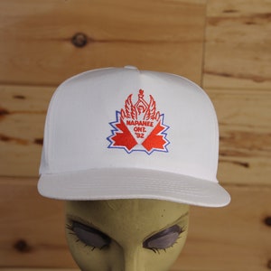Op de afbeelding: Witte baseballpet met een rood en blauw geborduurd logo met de tekst "Napanee Ont. '92".
