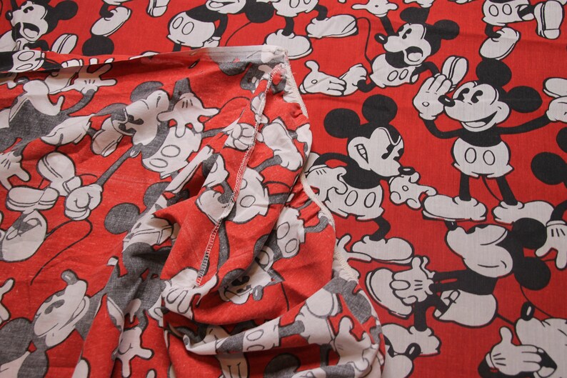 Op de afbeelding: Rode stof met een herhalend patroon van zwarte en witte Mickey Mouse-figuren. De stof is dubbelgevouwen, waardoor het patroon aan beide kanten zichtbaar is.