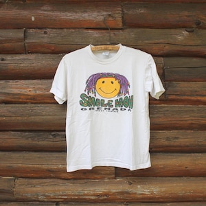 Może przedstawiać: Biała koszulka z grafiką przedstawiającą uśmiechniętą twarz z fioletowymi dredami oraz napisem "Smile Mon Grenada West Indies" w kolorze zielonym i żółtym. Koszulka wisi na drewnianym tle.