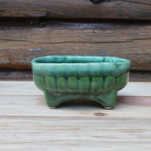 Peut inclure: Petit pot rectangulaire en céramique avec glaçure verte brillante. Le pot a trois petits pieds et un motif décoratif à nervures autour de la base. Le fond est une surface en bois.