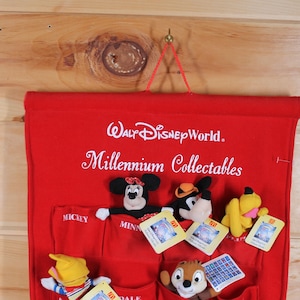 May include: Red felt wall hanging with the text "Walt Disney World. Millennium Collectibles" and 12 pockets for storing small items. The pockets are decorated with various Disney characters, including Mickey Mouse, Minnie Mouse, Donald Duck, and Goofy.