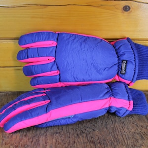 Puede incluir: Un par de guantes de invierno con una combinación de colores vibrantes. Los guantes son principalmente azules con acentos rosas brillantes en los dedos y a los lados. Los puños son de punto azul oscuro. La etiqueta dice "Thinsulate".