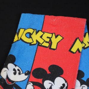 May include: A folded towel featuring Mickey Mouse. The towel is divided into blue and red sections, with the words "MICKEY MOUSE" in yellow. Mickey Mouse's face is visible on the blue section, and his body is on the red section.