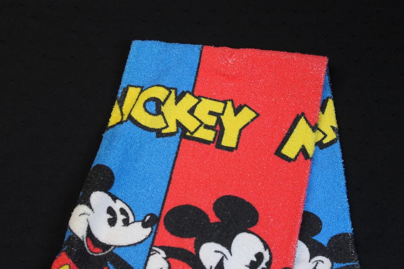 May include: A folded towel featuring Mickey Mouse. The towel is divided into blue and red sections, with the words "MICKEY MOUSE" in yellow. Mickey Mouse's face is visible on the blue section, and his body is on the red section.