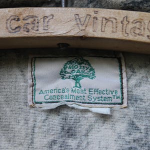 May include: A label with the text "Mossy Oak" and "America's Most Effective Concealment System" in green on a white background. The label is sewn onto camouflage fabric. A wooden hanger with the engraved text "rk car Vintage" is above the label.