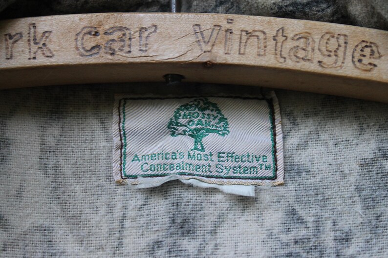 May include: A label with the text "Mossy Oak" and "America's Most Effective Concealment System" in green on a white background. The label is sewn onto camouflage fabric. A wooden hanger with the engraved text "rk car Vintage" is above the label.