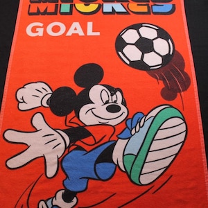 May include: Orange rectangular towel featuring Mickey Mouse kicking a soccer ball. The text "MICKEY GOAL" is at the top. Mickey is wearing a red shirt, blue pants, and blue and green shoes. The soccer ball is black and white.