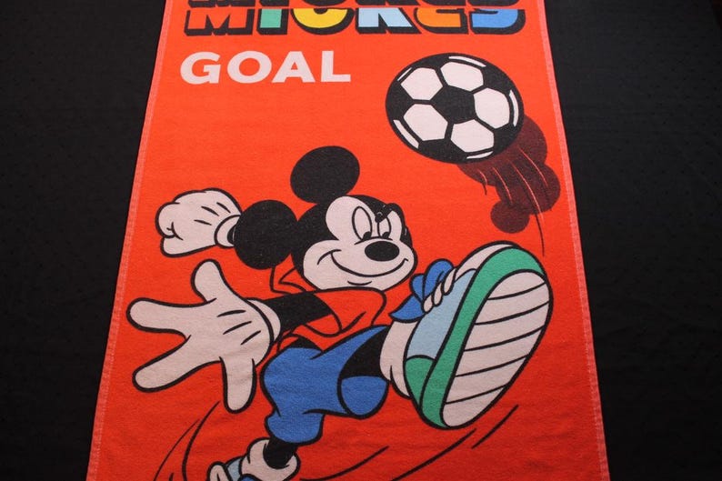 May include: Orange rectangular towel featuring Mickey Mouse kicking a soccer ball. The text "MICKEY GOAL" is at the top. Mickey is wearing a red shirt, blue pants, and blue and green shoes. The soccer ball is black and white.