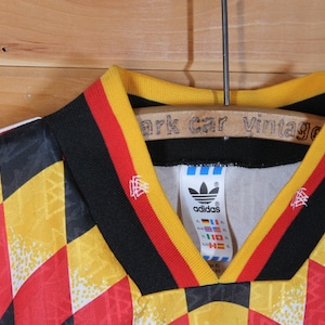 May include: A black, yellow, and red soccer jersey with a checkered pattern. The jersey has a black collar with yellow and red stripes. The Adidas logo is on the left chest with the word "adidas" underneath. The jersey has a white tag with the Adidas logo and the words "adidas" and "Germany" on it.