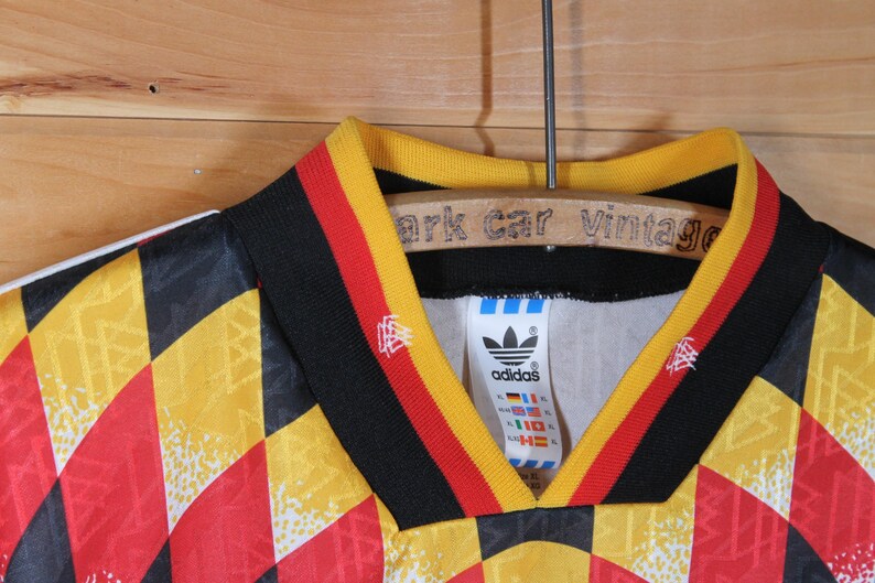 May include: A black, yellow, and red soccer jersey with a checkered pattern. The jersey has a black collar with yellow and red stripes. The Adidas logo is on the left chest with the word "adidas" underneath. The jersey has a white tag with the Adidas logo and the words "adidas" and "Germany" on it.