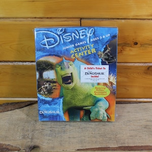 May include: A Disney Junior Games Activity Center box featuring a green dinosaur illustration with an open mouth. The box has a blue background with the text "DINOSAUR" and "AGES 5 & UP". Includes "A Child's Ticket To Dinosaur Inside!"