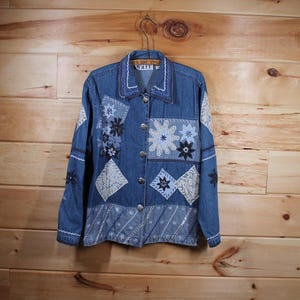 May include: A blue denim jacket with a patchwork design featuring embroidered flowers and geometric shapes. The jacket has a button closure and a collar.