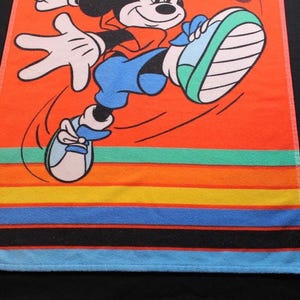 May include: Orange beach towel featuring a cartoon Mickey Mouse in a running pose. The towel has colorful horizontal stripes in shades of orange, yellow, green, blue, and black. The towel is made of a soft, absorbent material.