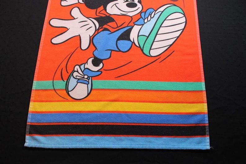May include: Orange beach towel featuring a cartoon Mickey Mouse in a running pose. The towel has colorful horizontal stripes in shades of orange, yellow, green, blue, and black. The towel is made of a soft, absorbent material.