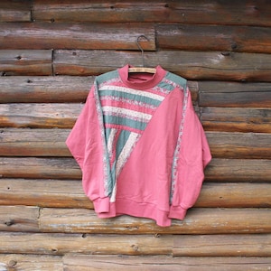 May include: A vintage pink sweatshirt with a high neckline and long sleeves. The front features a geometric design with stripes of white, green, and pink. The sweatshirt hangs on a metal hanger against a wooden log background.