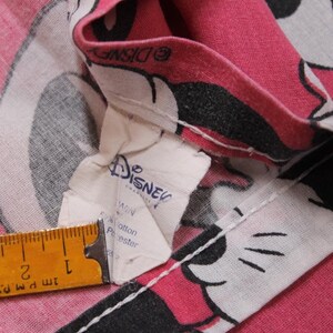 Peut inclure: Gros plan sur un tissu rose avec un motif de personnage de dessin anim&eacute;. Le tissu pr&eacute;sente une impression de personnage blanc, noir et rose. Une &eacute;tiquette Disney est visible, ainsi qu'une r&egrave;gle montrant des mesures en cm.