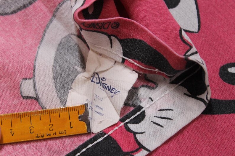 Peut inclure: Gros plan sur un tissu rose avec un motif de personnage de dessin anim&eacute;. Le tissu pr&eacute;sente une impression de personnage blanc, noir et rose. Une &eacute;tiquette Disney est visible, ainsi qu'une r&egrave;gle montrant des mesures en cm.