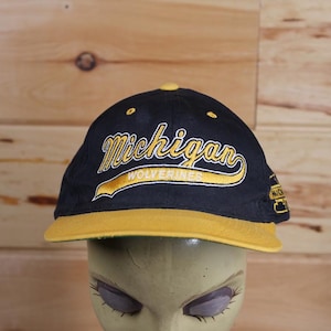 Op de afbeelding: Een zwarte en gouden baseballpet met de tekst "Michigan Wolverines" geborduurd op de voorkant.
