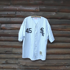 May include: White baseball jersey with black pinstripes, the number 45, and the Chicago White Sox logo. The jersey is hanging on a wooden hanger against a rustic wooden background.