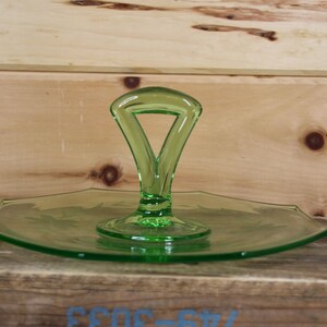 Op de afbeelding: Een groene glazen serveerschaal met een handvat. De schaal heeft een driehoekig handvat en een ronde basis. De schaal is lichtgroen van kleur en heeft een bloemenontwerp dat in het glas is geëtst. De schaal staat op een houten oppervlak.