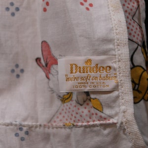 May include: A white cotton baby blanket with a pink and blue polka dot pattern. The blanket features a cartoon image of a duck and a dog. The label reads "Dundee "we're soft on babies" made in U.S.A. 100% cotton."
