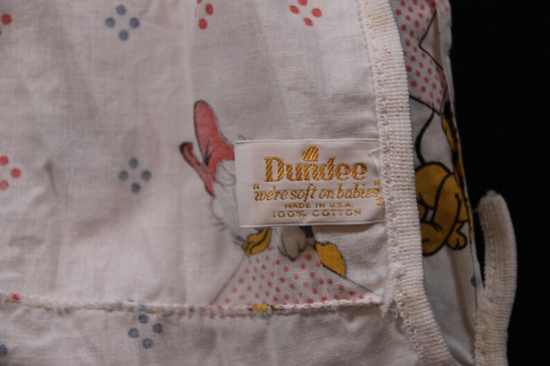 May include: A white cotton baby blanket with a pink and blue polka dot pattern. The blanket features a cartoon image of a duck and a dog. The label reads "Dundee "we're soft on babies" made in U.S.A. 100% cotton."