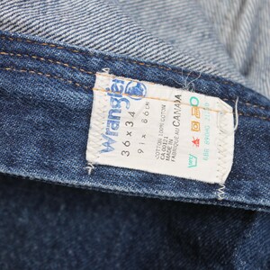 May include: A blue denim Wrangler jeans label with white text. The label reads "Wrangler", "36 x 34", "91 x 86 cm", "CA 00121", "Cotton 100% Coton", "Made in", "Fabrique au Canada", "6BR 890HG 21:30".