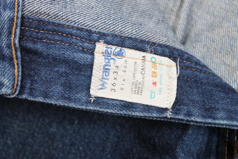 May include: A blue denim Wrangler jeans label with white text. The label reads "Wrangler", "36 x 34", "91 x 86 cm", "CA 00121", "Cotton 100% Coton", "Made in", "Fabrique au Canada", "6BR 890HG 21:30".
