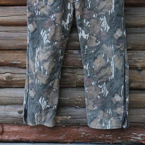 Puede incluir: Pantalones de camuflaje con un estampado de hojas en tonos marrones, grises y blancos. Los pantalones tienen una cremallera en el lateral de cada pierna. El fondo es una pared de madera.