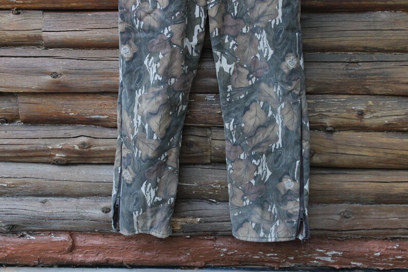 Puede incluir: Pantalones de camuflaje con un estampado de hojas en tonos marrones, grises y blancos. Los pantalones tienen una cremallera en el lateral de cada pierna. El fondo es una pared de madera.
