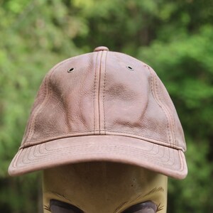 Puede incluir: Una gorra de béisbol de cuero marrón con una visera curvada y dos pequeños ojales en la parte superior.