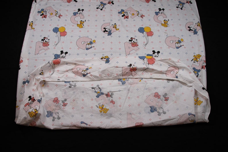 May include: A white fitted sheet with a pink and blue Mickey Mouse and Minnie Mouse print. The sheet features a repeating pattern of Mickey and Minnie Mouse characters holding balloons and letters of the alphabet.