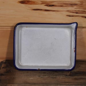May include: A white enamel baking dish with a blue rim. The dish is rectangular and has a slightly rounded edge. It is sitting on a wooden surface.
