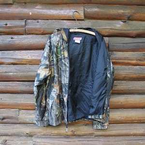 May include: Camouflage pattern jacket with a black interior lining. The jacket is hanging on a wooden hanger against a backdrop of wooden logs. The jacket features a zipper closure and a hood.