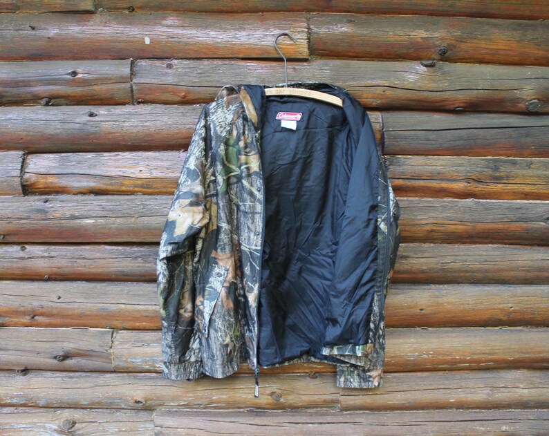 May include: Camouflage pattern jacket with a black interior lining. The jacket is hanging on a wooden hanger against a backdrop of wooden logs. The jacket features a zipper closure and a hood.
