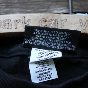 May include: A black t-shirt featuring a rectangular black fabric tag with white text detailing Spider-Man 3 copyright information from 2007. Below it, a white care instruction label lists 100% cotton material and manufacturing details.