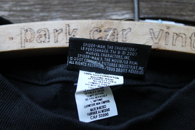 May include: A black t-shirt featuring a rectangular black fabric tag with white text detailing Spider-Man 3 copyright information from 2007. Below it, a white care instruction label lists 100% cotton material and manufacturing details.