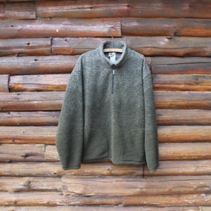 May include: A green fleece pullover with a quarter-zip collar hangs on a wooden hanger against a backdrop of wooden logs. The pullover has a textured appearance and a dark zipper.
