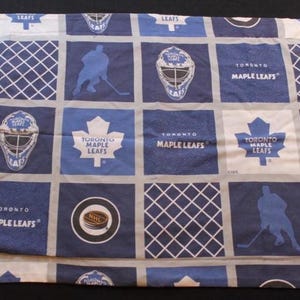 May include: A blue and white pillowcase featuring a repeating pattern of hockey-related imagery. The design includes hockey players, goalie masks, the Toronto Maple Leafs logo, and the NHL logo. The pillowcase is made of a soft fabric.