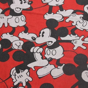 Op de afbeelding: Rode stof met een herhalend patroon van zwarte en witte Mickey Mouse-figuren. De figuren staan in verschillende poses en hebben een cartoonachtige stijl.