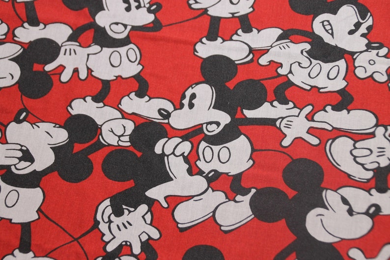 Op de afbeelding: Rode stof met een herhalend patroon van zwarte en witte Mickey Mouse-figuren. De figuren staan in verschillende poses en hebben een cartoonachtige stijl.