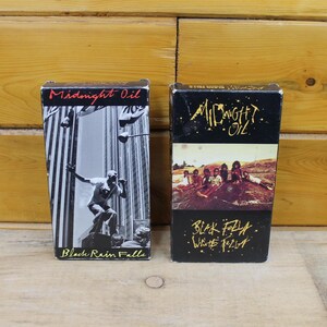 Puede incluir: Dos cintas de cassette vintage de Midnight Oil. Una presenta una imagen en blanco y negro de una persona cantando con el texto "Black Rain Falls". La otra tiene una foto de la banda con el texto "Black Fall White Fall."